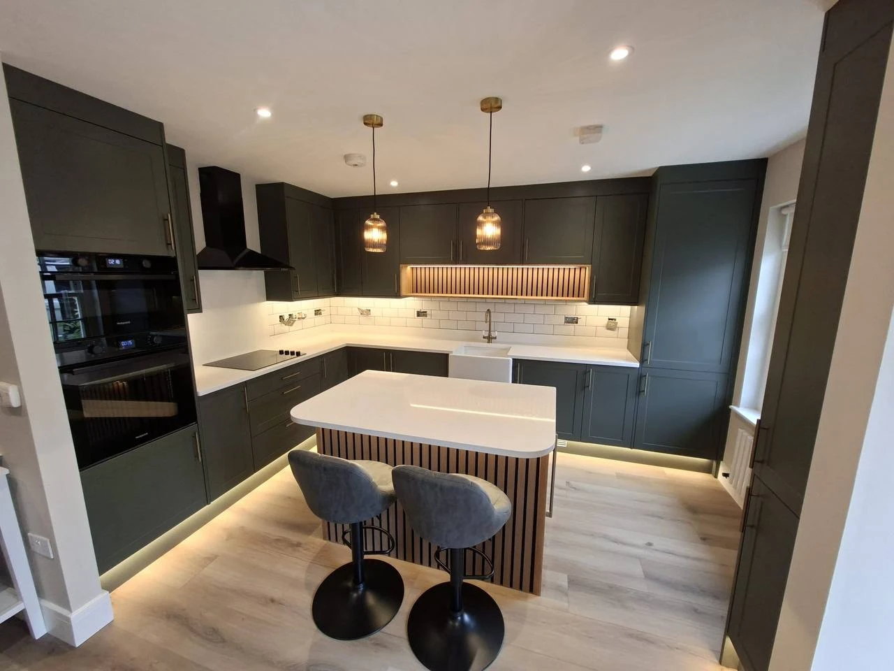 Dark green shaker kitchen with an oak-slatted island, white worktops and brass pendant lights